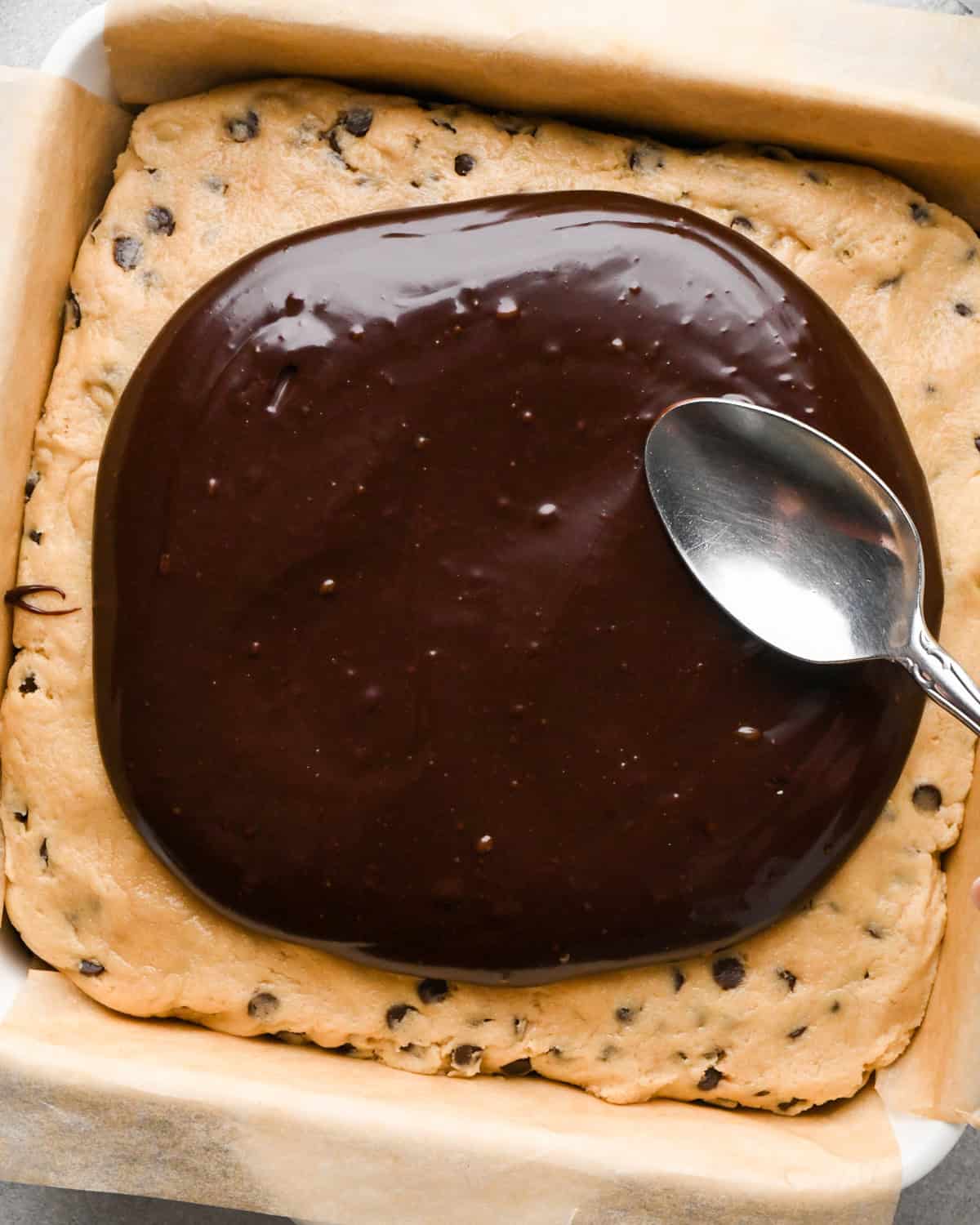 assembling cookie dough brownies - a silver spoon beginning to spread chocolate ganache over the cookie dough layer