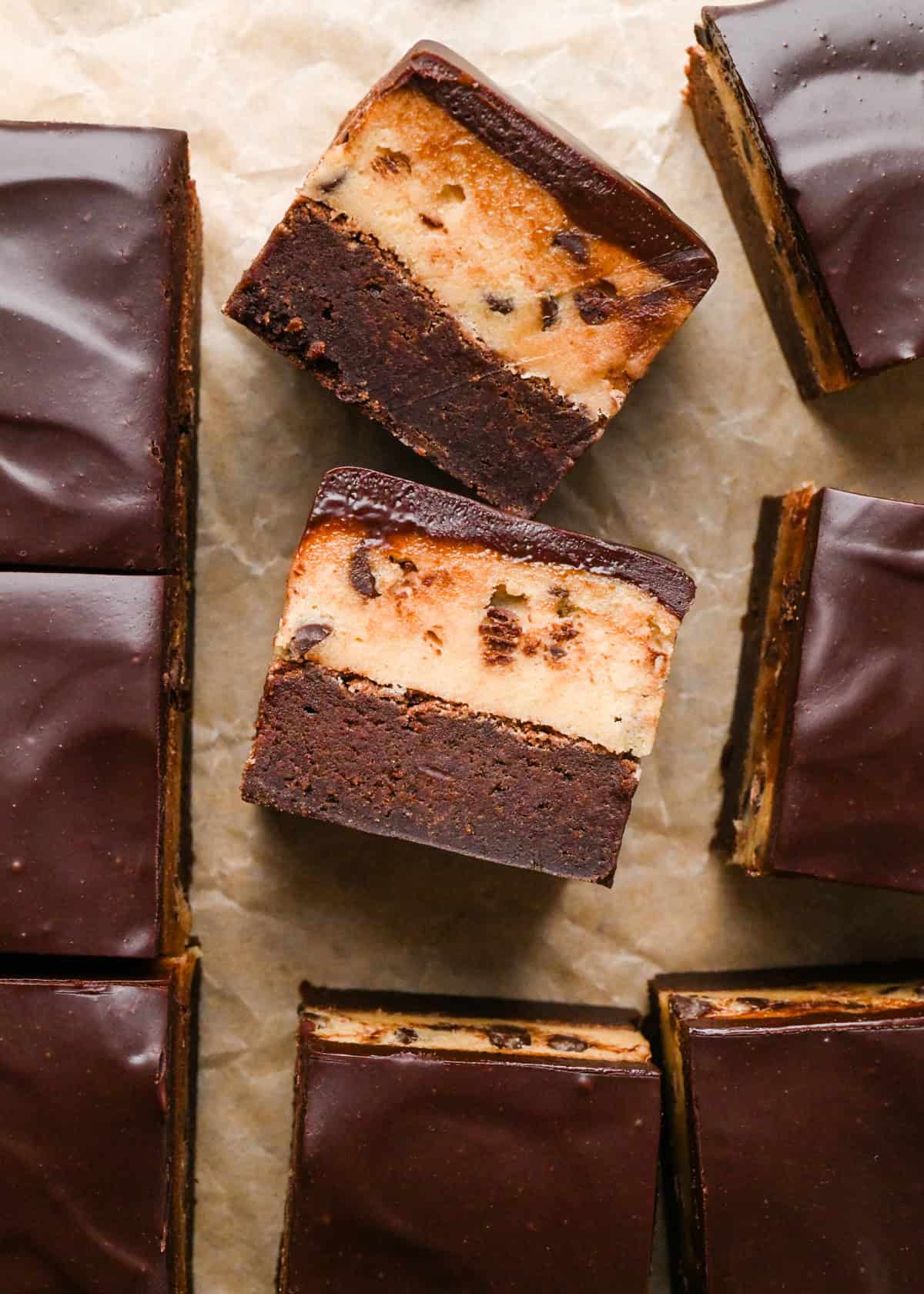 two cookie dough brownies turned on their side with a fudgy chocolate brownie on the bottom, edible chocolate chip cookie dough in the middle, and a glossy chocolate ganache on top on parchment paper. Surrounded by other cookie dough brownies.