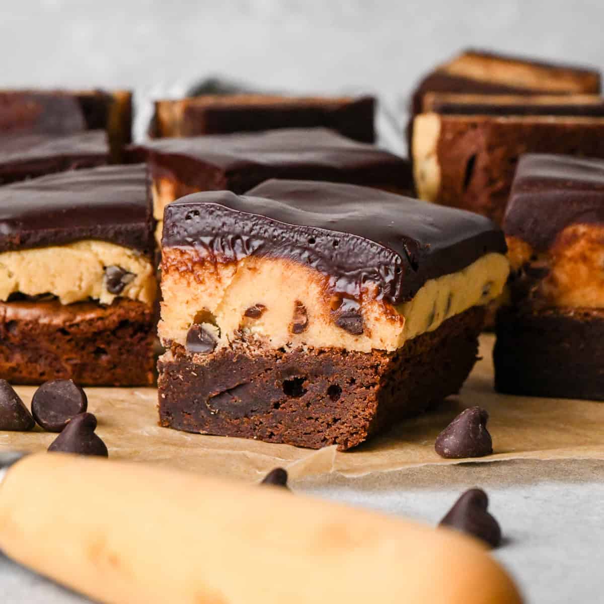 3 cookie dough brownies with a fudgy chocolate brownie on the bottom, edible chocolate chip cookie dough in the middle, and a glossy chocolate ganache on top on parchment paper. Surrounded by other cookie dough brownies and chocolate chips