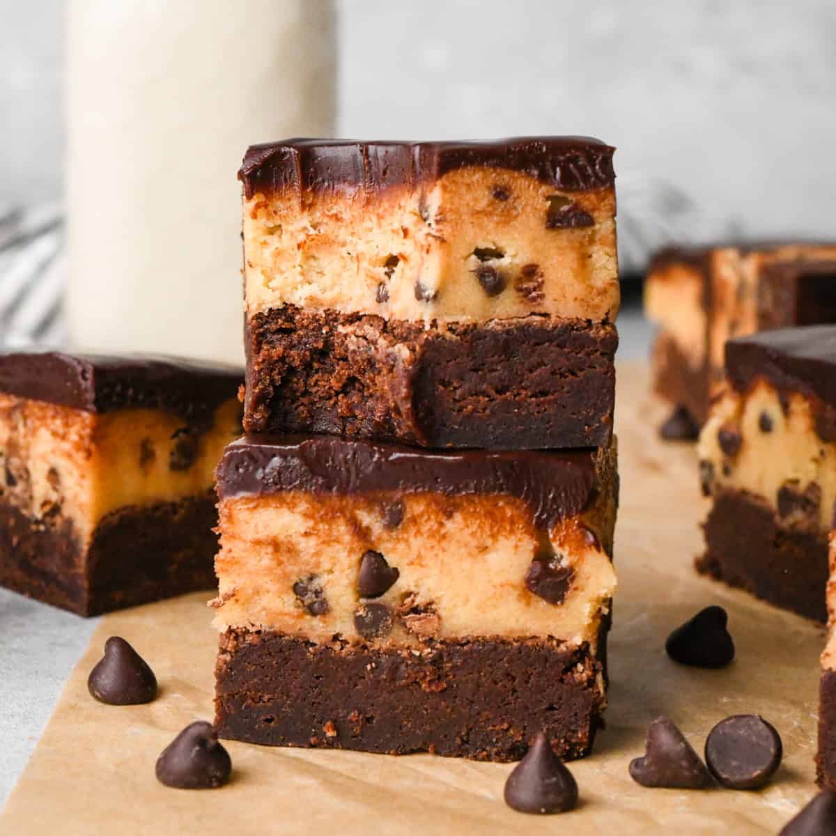 a stack of two cookie dough brownies with a fudgy chocolate brownie on the bottom, edible chocolate chip cookie dough in the middle, and a glossy chocolate ganache on top on parchment paper. Surrounded by other cookie dough brownies and chocolate chips