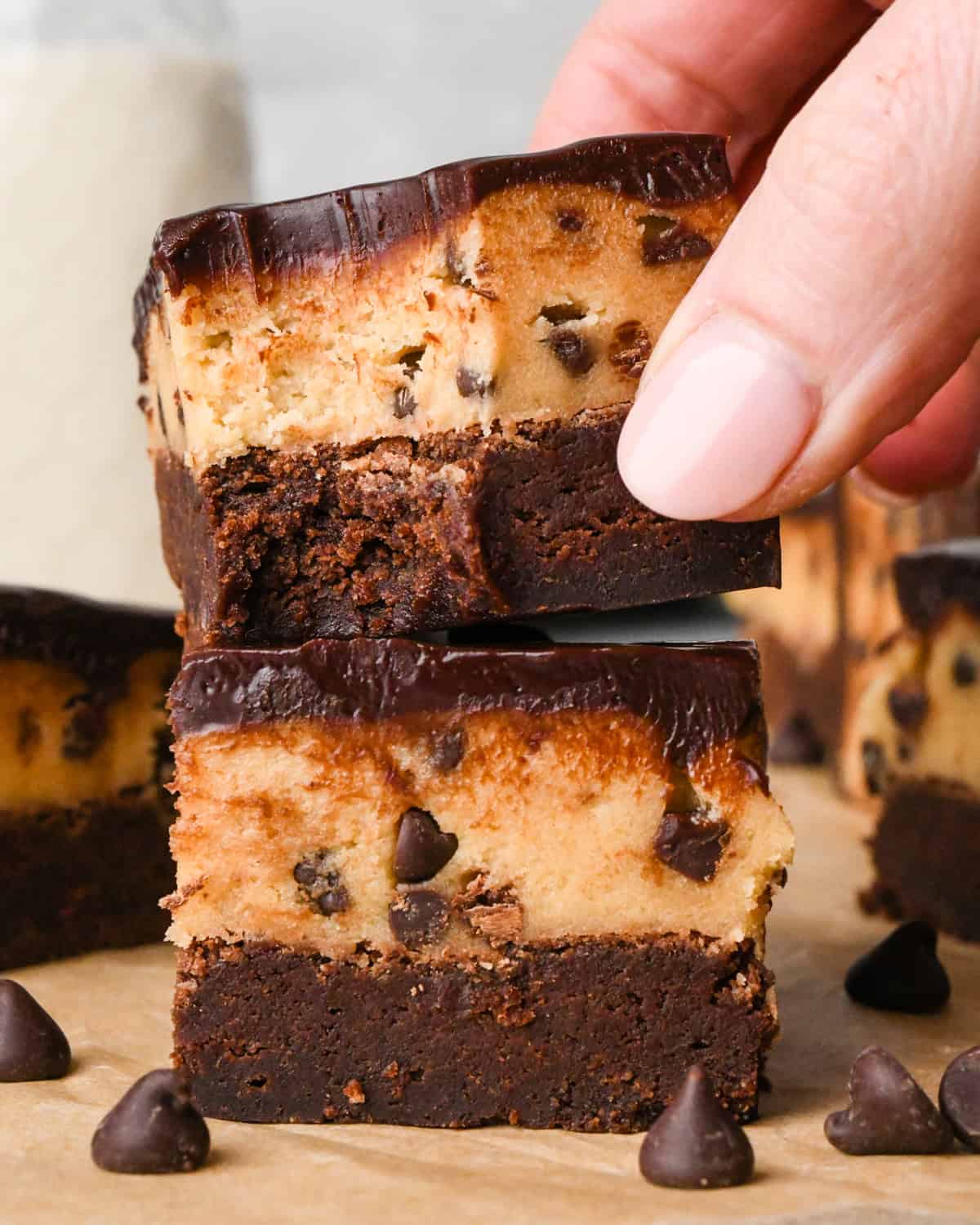 a stack of two cookie dough brownies with a fudgy chocolate brownie on the bottom, edible chocolate chip cookie dough in the middle, and a glossy chocolate ganache on top on parchment paper A hand is picking up the top brownie which has a bite taken out of it. Surrounded by other cookie dough brownies and chocolate chips