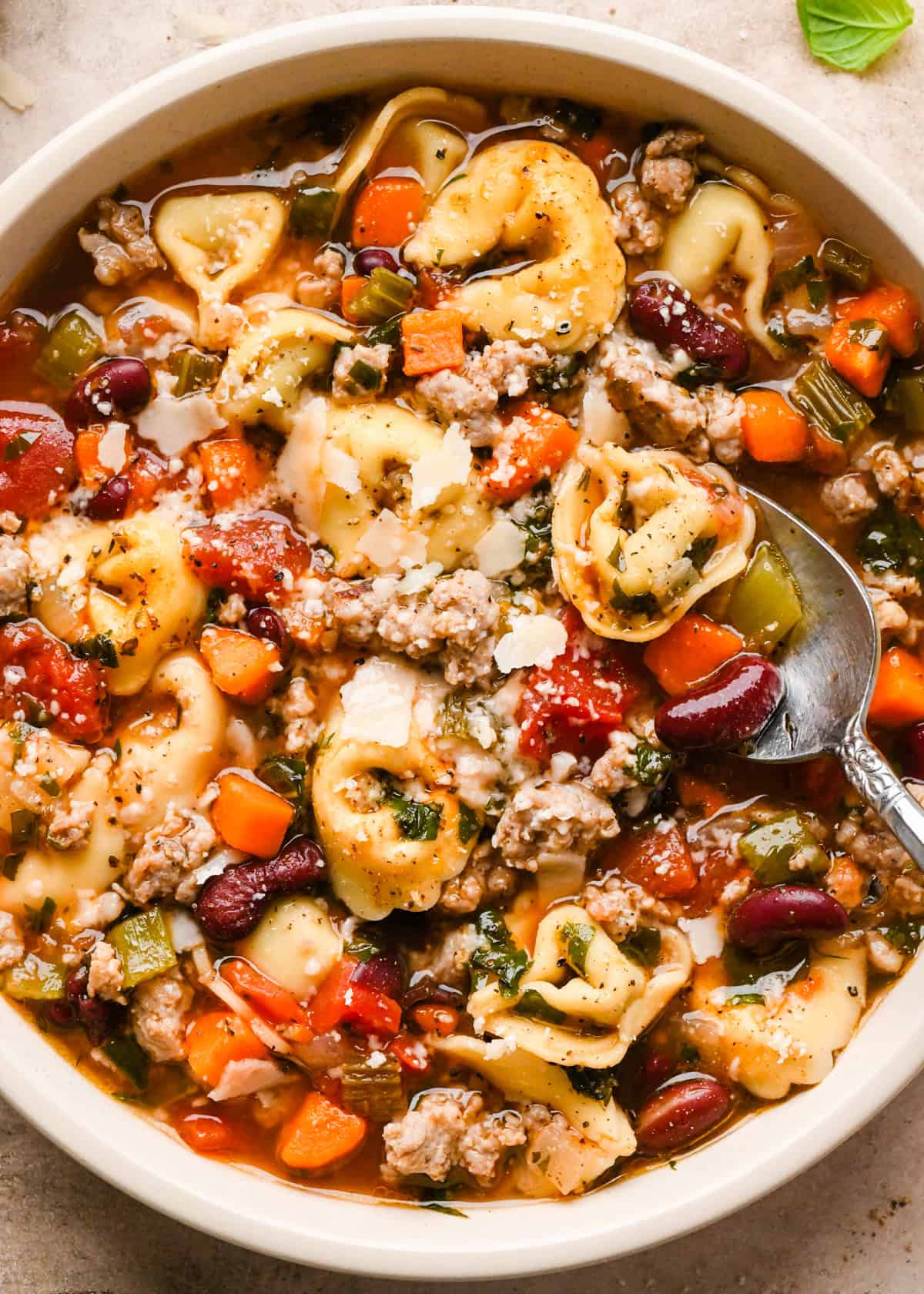 A white bowl of sausage tortellini soup with cheese-filled tortellini, crumbled sausage, carrots, celery, beans, and herbs in a rich tomato broth, with a spoon resting inside the bowl.