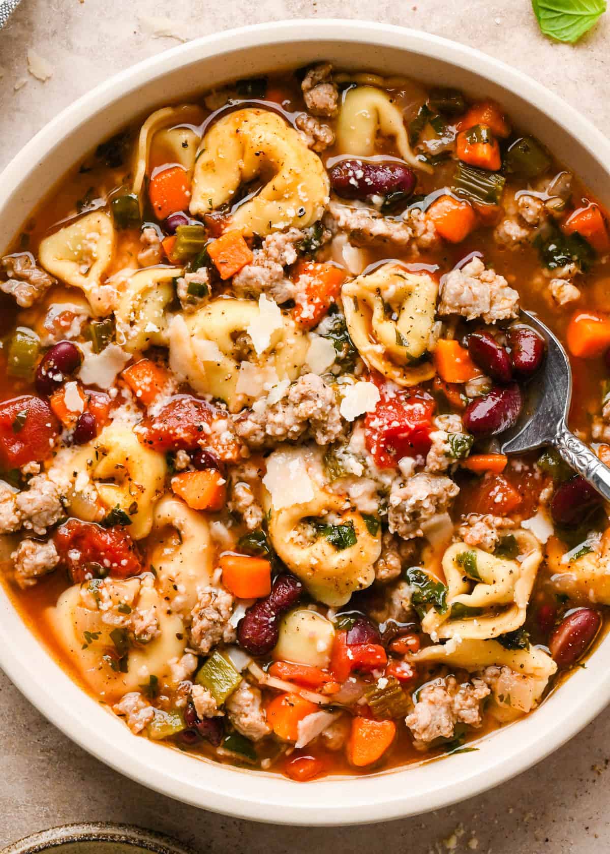 A white bowl of sausage tortellini soup with cheese-filled tortellini, crumbled sausage, carrots, celery, beans, and herbs in a rich tomato broth, with a spoon resting inside the bowl.