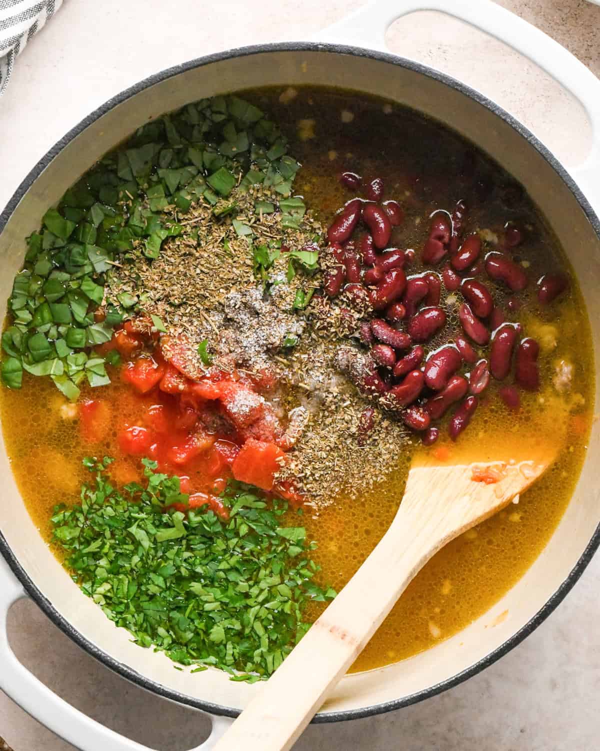 Making sausage tortellini soup: overhead photo of chicken broth, beans, tomatoes, spices, and herbs added to a large pot along with the cooked meat and vegetables.