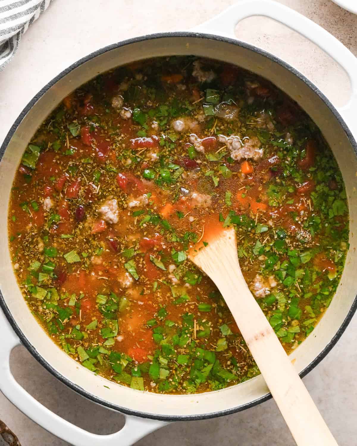 Making sausage tortellini soup: overhead photo of chicken broth, beans, tomatoes, spices, and herbs added to a large pot along with the cooked meat and vegetables and being stirred together with a wooden spoon.