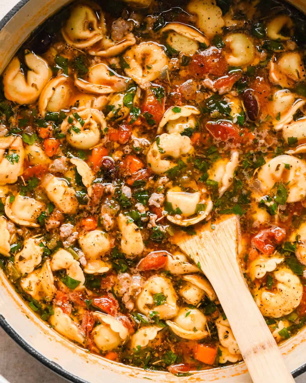 Close-up photo of a pot of sausage tortellini soup with cheese-filled tortellini, crumbled sausage, carrots, celery, beans, and herbs in a rich tomato broth, being stirred together with a wooden spoon.