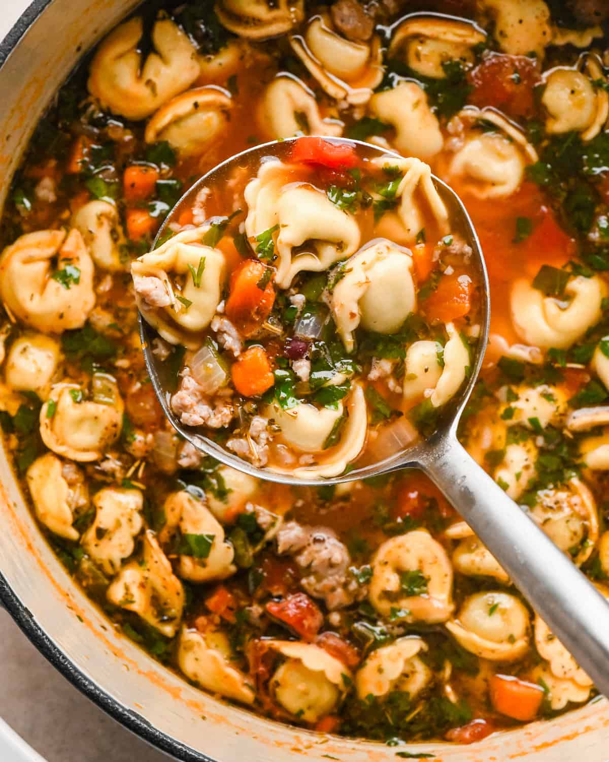 Close-up photo of a pot of sausage tortellini soup with cheese-filled tortellini, crumbled sausage, carrots, celery, beans, and herbs in a rich tomato broth, being scooped with a ladle.