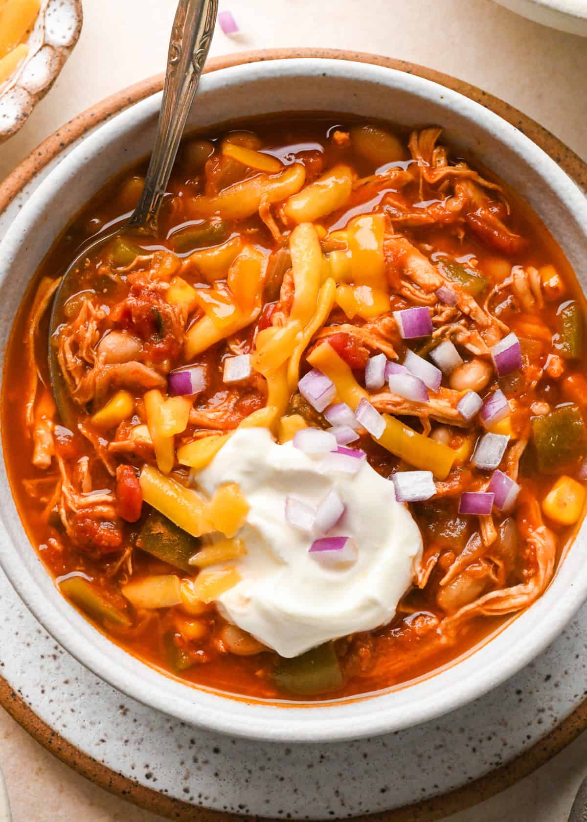 a bowl of chicken chili with a spoon topped with sour cream, red onions and cheese 