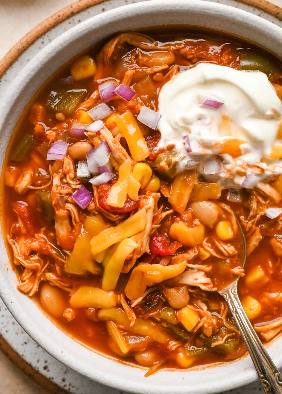 a spoon taking a scoop out of a bowl of Slow Cooker Chicken Chili topped with shredded cheese, sour cream and red onion. 