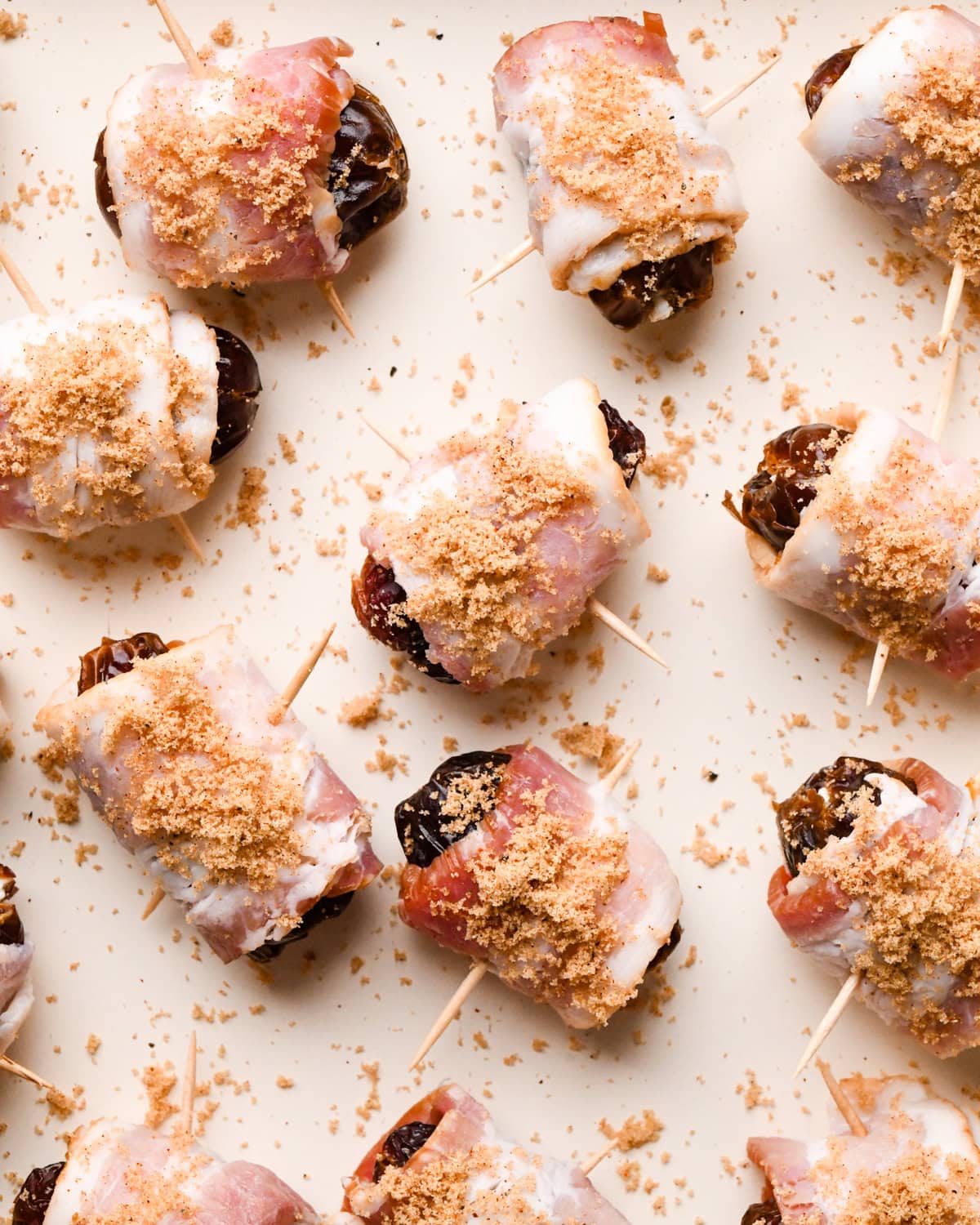 Making bacon-wrapped dates: close-up photo of bacon-wrapped dates topped with brown sugar topping and arranged on a light-colored cooking sheet.