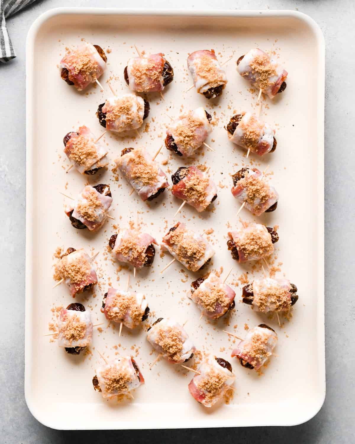Making bacon-wrapped dates: overhead photo of bacon-wrapped dates topped with brown sugar topping and arranged on a light-colored cooking sheet.