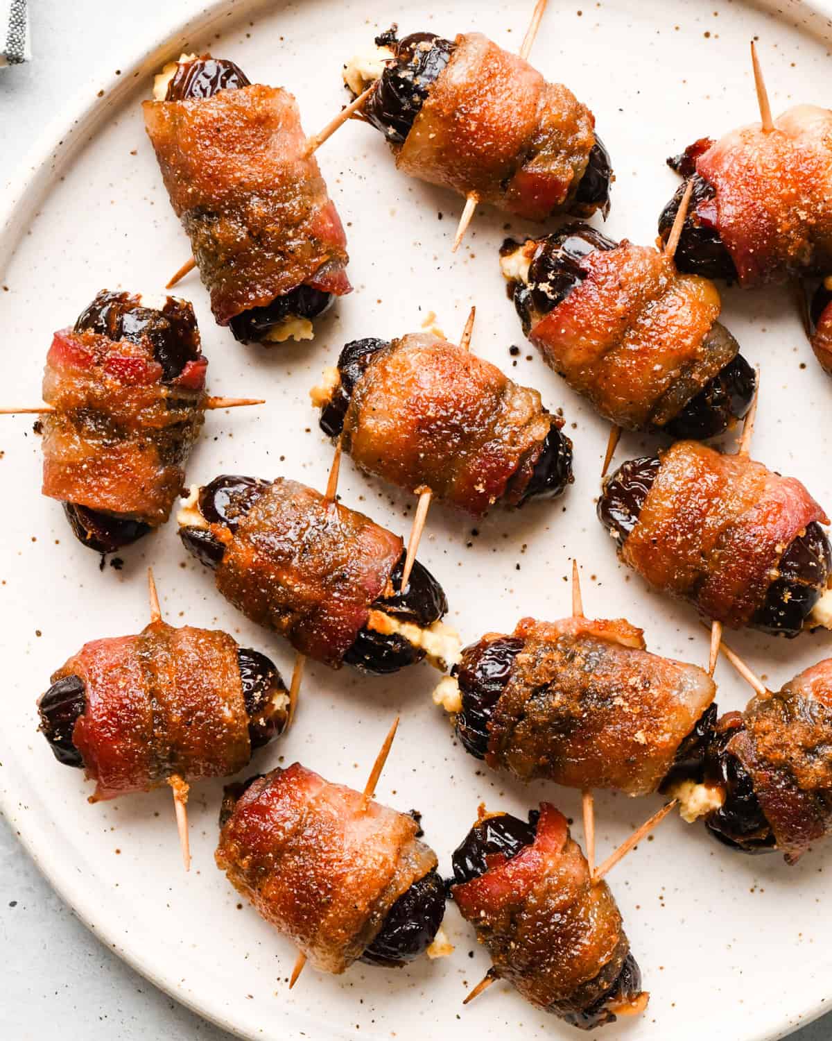 Close-up of bacon-wrapped dates secured with toothpicks, baked until crispy and glazed, arranged on a light-colored plate.