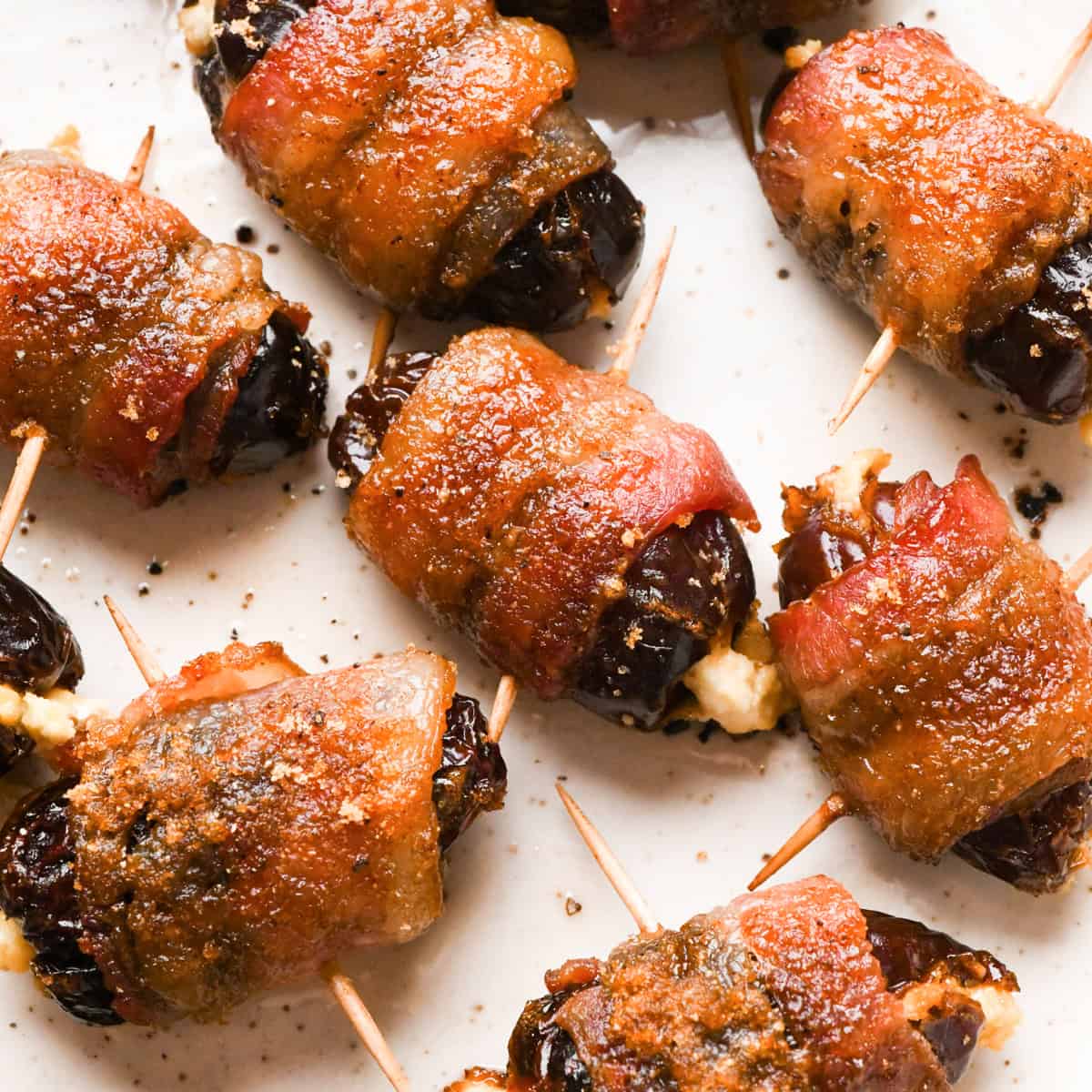 Close-up of bacon-wrapped dates secured with toothpicks, baked until crispy and glazed, arranged on a light-colored plate.