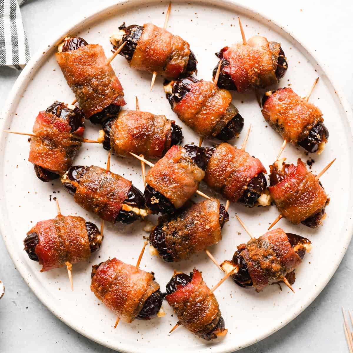 Close-up of bacon-wrapped dates secured with toothpicks, baked until crispy and glazed, arranged on a light-colored plate.