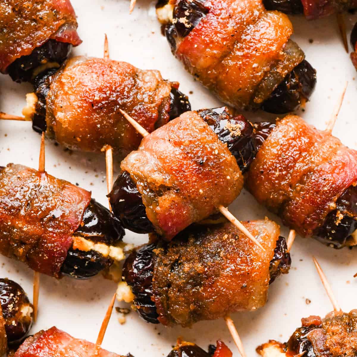 Close-up of bacon-wrapped dates secured with toothpicks, baked until crispy and glazed, arranged on a light surface.