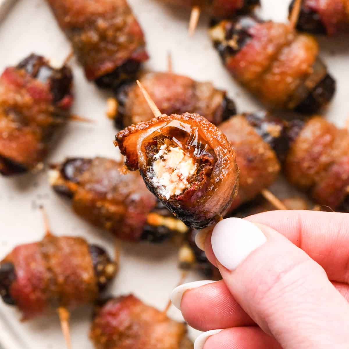 Close-up of bacon-wrapped dates secured with toothpicks, baked until crispy and glazed, arranged on a light surface. A bite is taken out of one of the dates.