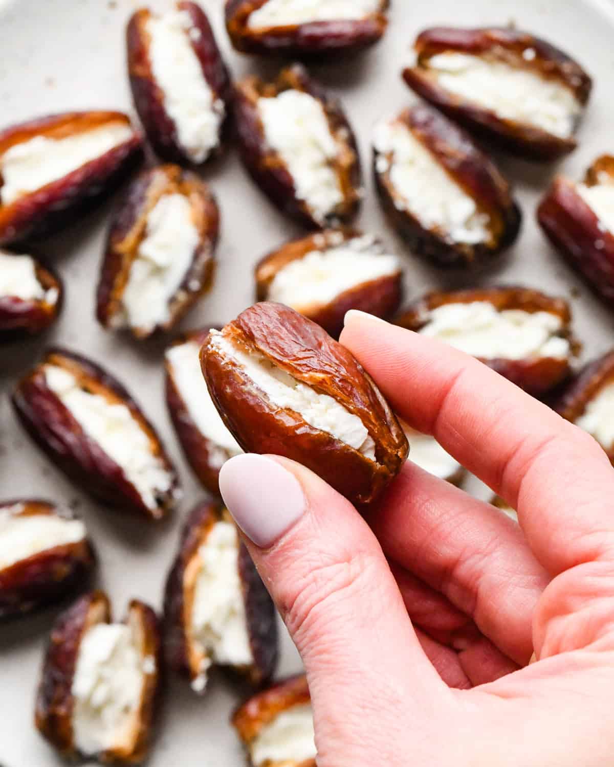 Making bacon-wrapped dates: overhead photo of a hand holding a goat-cheese filled date. A plate full of more stuffed dates is visible in the background.