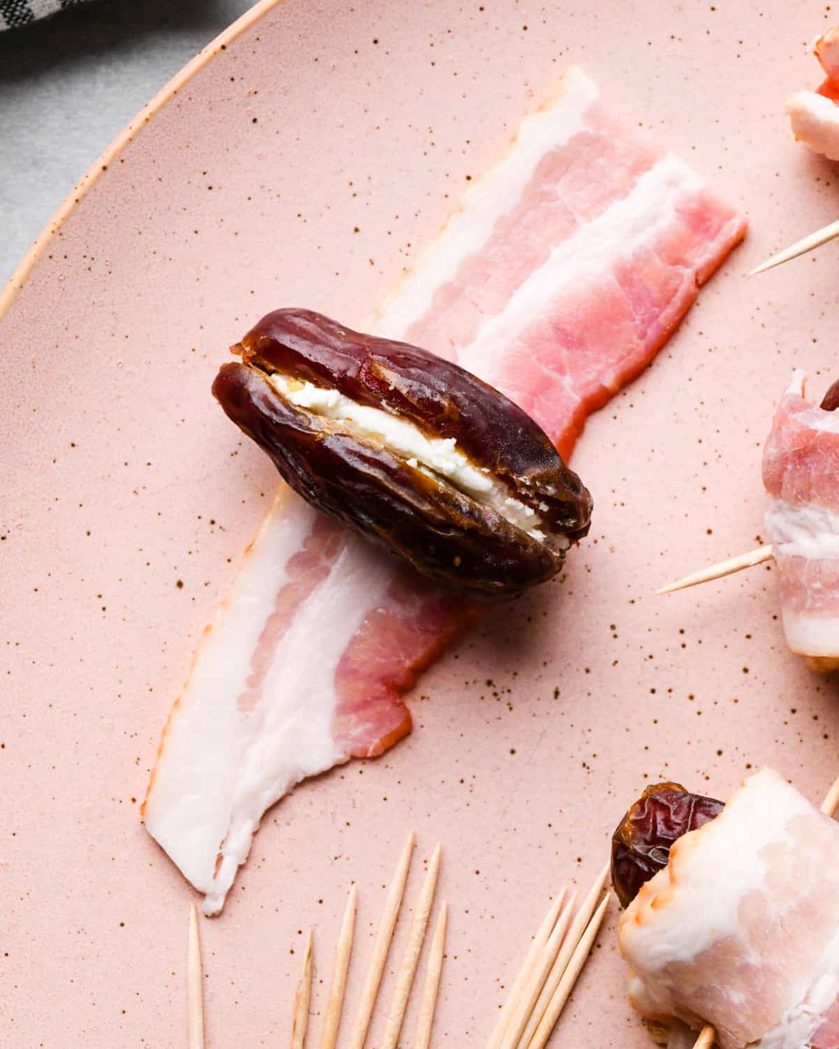 Making bacon-wrapped dates: overhead photo of a goat-cheese-stuffed date sitting on top of a strip of bacon ready to be wrapped and secured with a toothpick. More dates are surrounding it on a speckled plate.