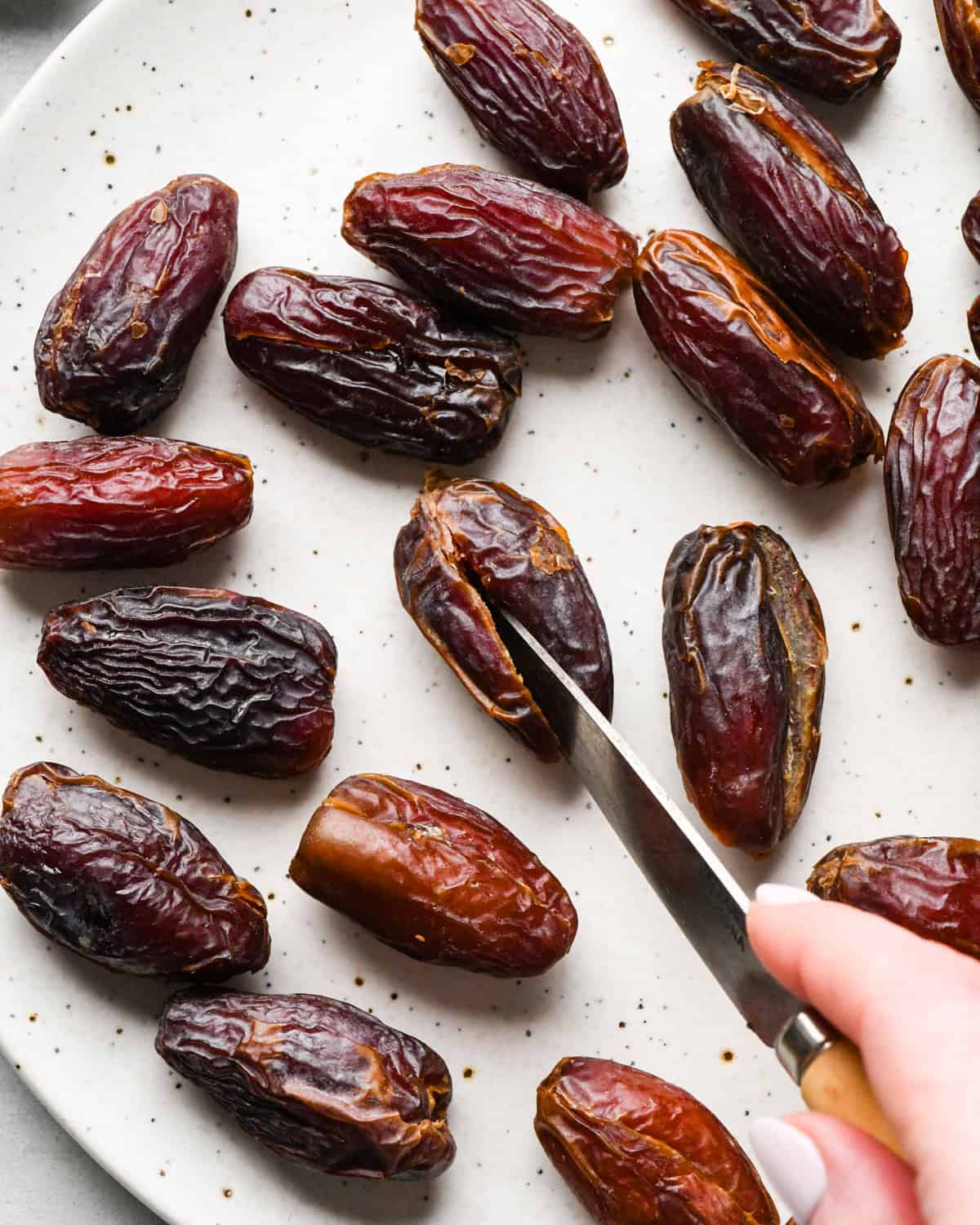 Making bacon-wrapped dates: overhead photo of dates arranged on a white speckled plate with a person cutting into one of the dates with a knife.