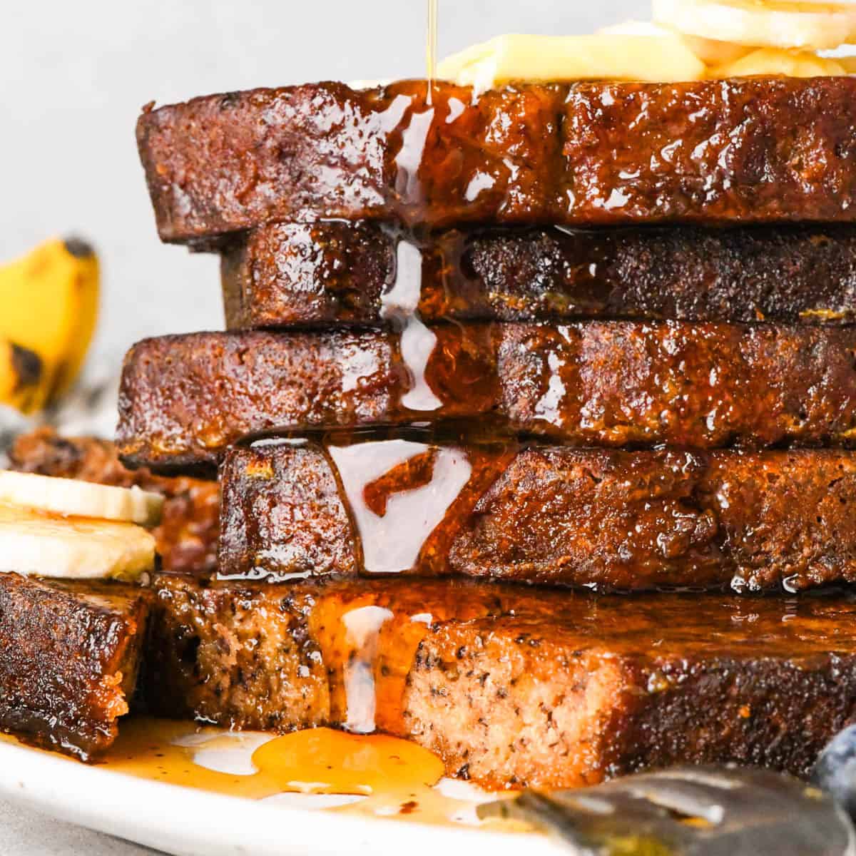 Close-up side view of a tall stack of banana bread French toast on a white plate, topped with butter and banana slices as maple syrup is poured over the top and drips down the golden-brown slices, with blueberries and extra banana slices blurred in the background.