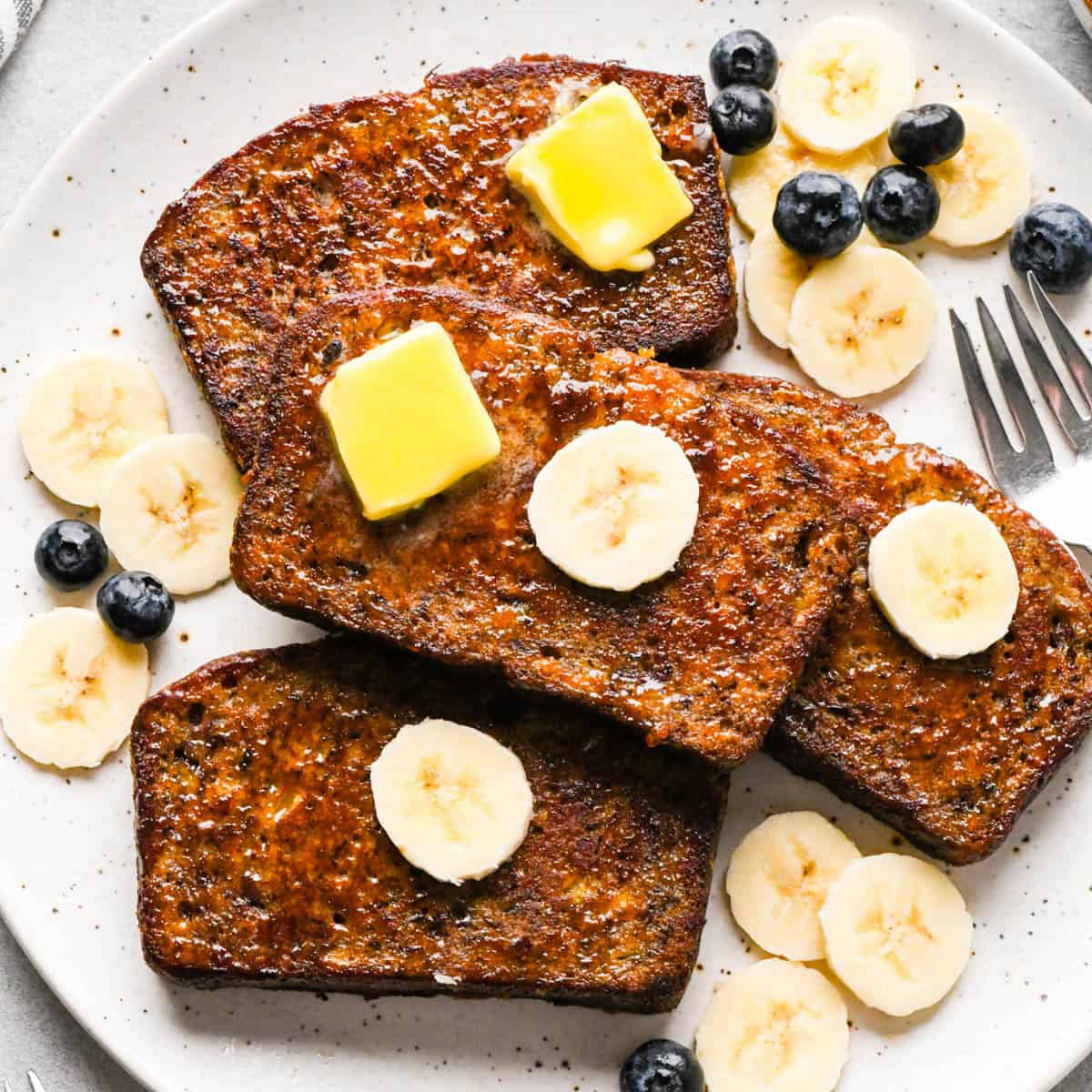 Overhead photo of four slices of golden-brown banana bread French toast on a white speckled plate, topped with melting pats of butter and banana slices, lightly dusted with powdered sugar and syrup, and served with fresh blueberries and extra banana slices on the side next to a fork.