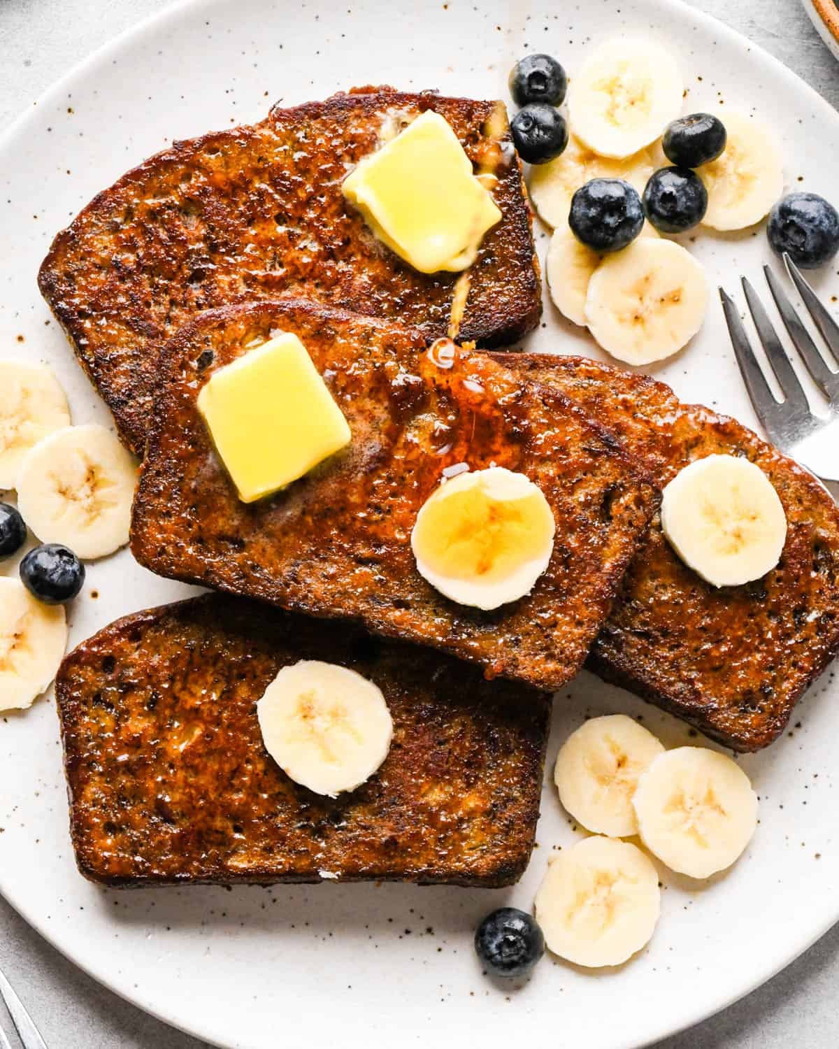Overhead photo of four slices of golden-brown banana bread French toast on a white speckled plate, topped with melting pats of butter and banana slices, lightly dusted with powdered sugar and syrup, and served with fresh blueberries and extra banana slices on the side next to a fork.