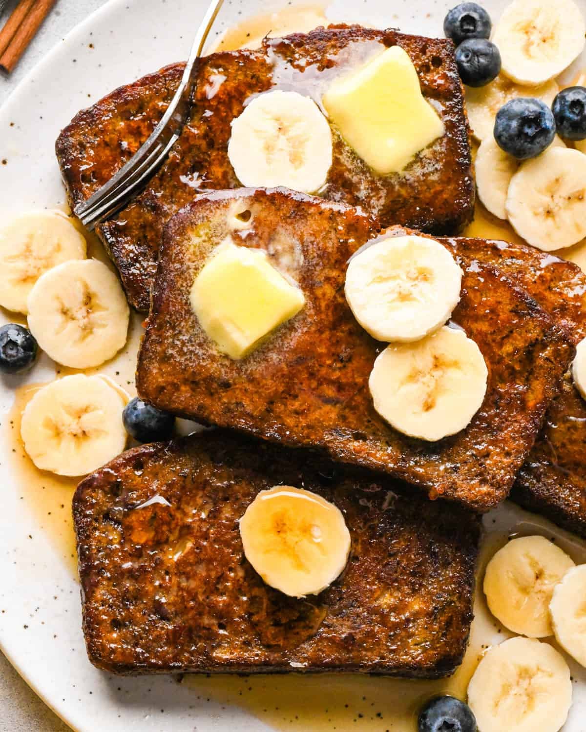 Overhead photo of four slices of golden-brown banana bread French toast on a white speckled plate, topped with melting pats of butter and banana slices, lightly dusted with powdered sugar and syrup, and served with fresh blueberries and extra banana slices on the side next to a fork.