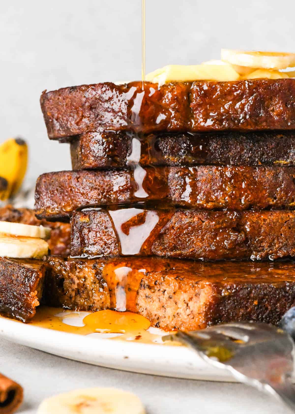 Close-up side view of a tall stack of banana bread French toast on a white plate, topped with butter and banana slices as maple syrup is poured over the top and drips down the golden-brown slices, with blueberries and extra banana slices blurred in the background.