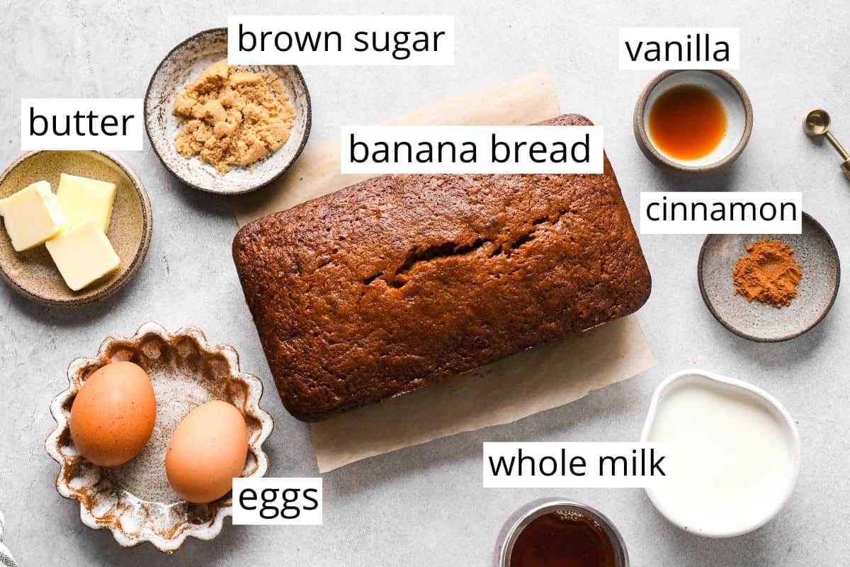 Overhead photo of the ingredients for banana bread French toast arranged on a light countertop, including a loaf of banana bread, eggs, butter, brown sugar, whole milk, vanilla, and cinnamon in small bowls with labels identifying each ingredient.