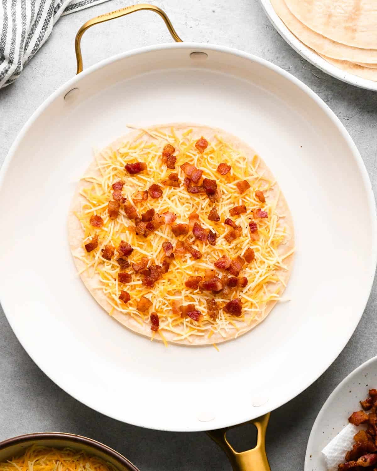 making breakfast quesadillas - a tortilla topped with Mexican blend cheese and bacon in a white skillet