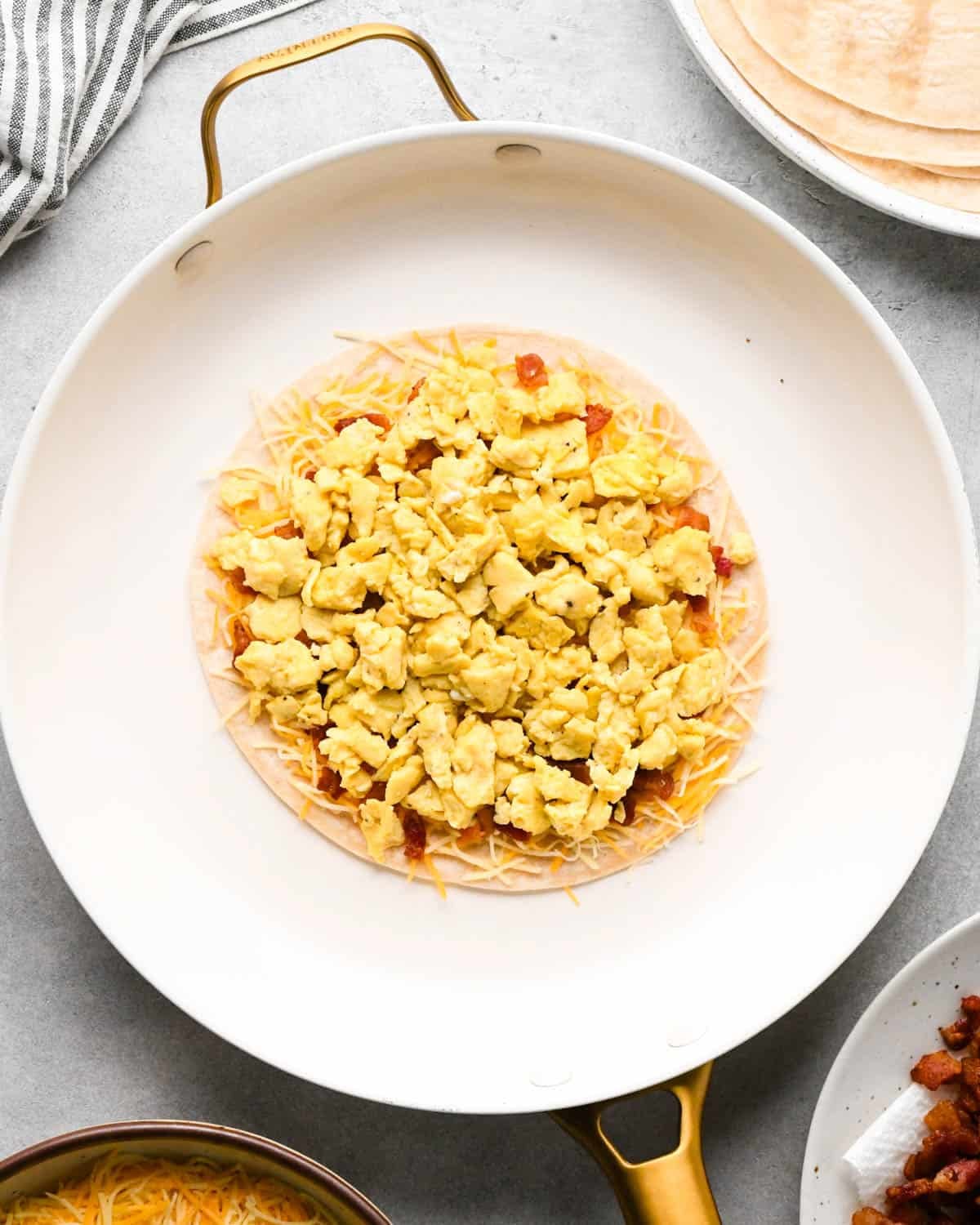 making breakfast quesadillas - a tortilla topped with Mexican blend cheese, bacon and scrambled eggs in a white skillet