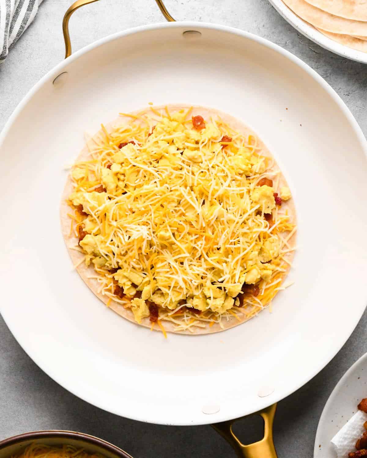 making breakfast quesadillas - cheese on top of eggs and bacon on a tortilla in a white skillet