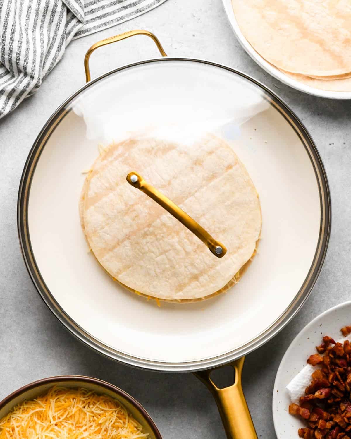 making breakfast quesadillas - an assembled breakfast quesadilla cooking in a white skillet with a clear glass lid on top