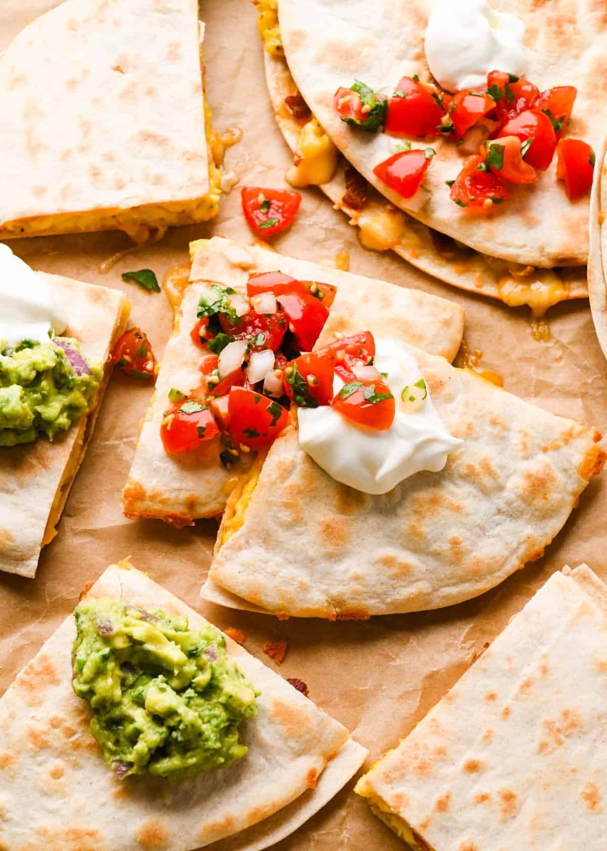breakfast quesadillas on parchment paper cut into triangles topped with sour cream, pico de gallo and guacamole