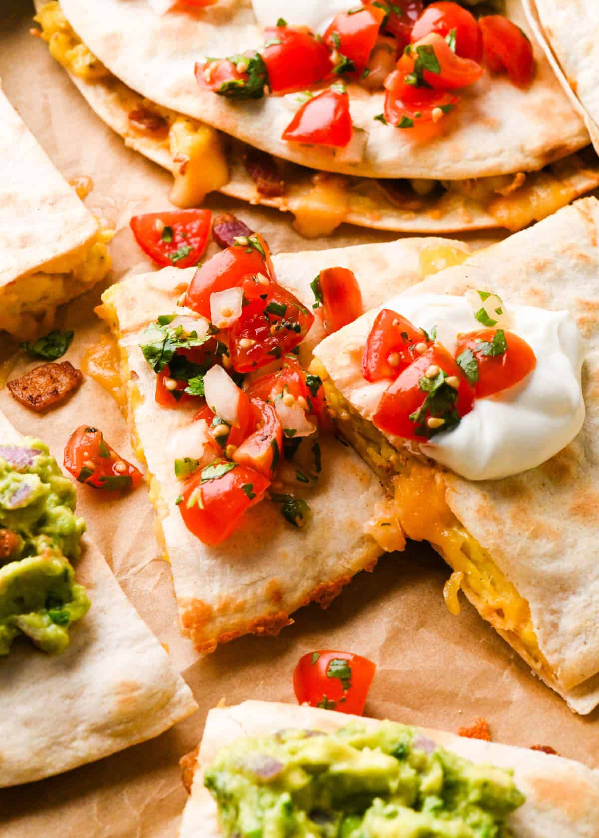 close up photo of breakfast quesadillas on parchment paper cut into triangles topped with sour cream, pico de gallo and guacamole the melty cheese and eggs inside are visible
