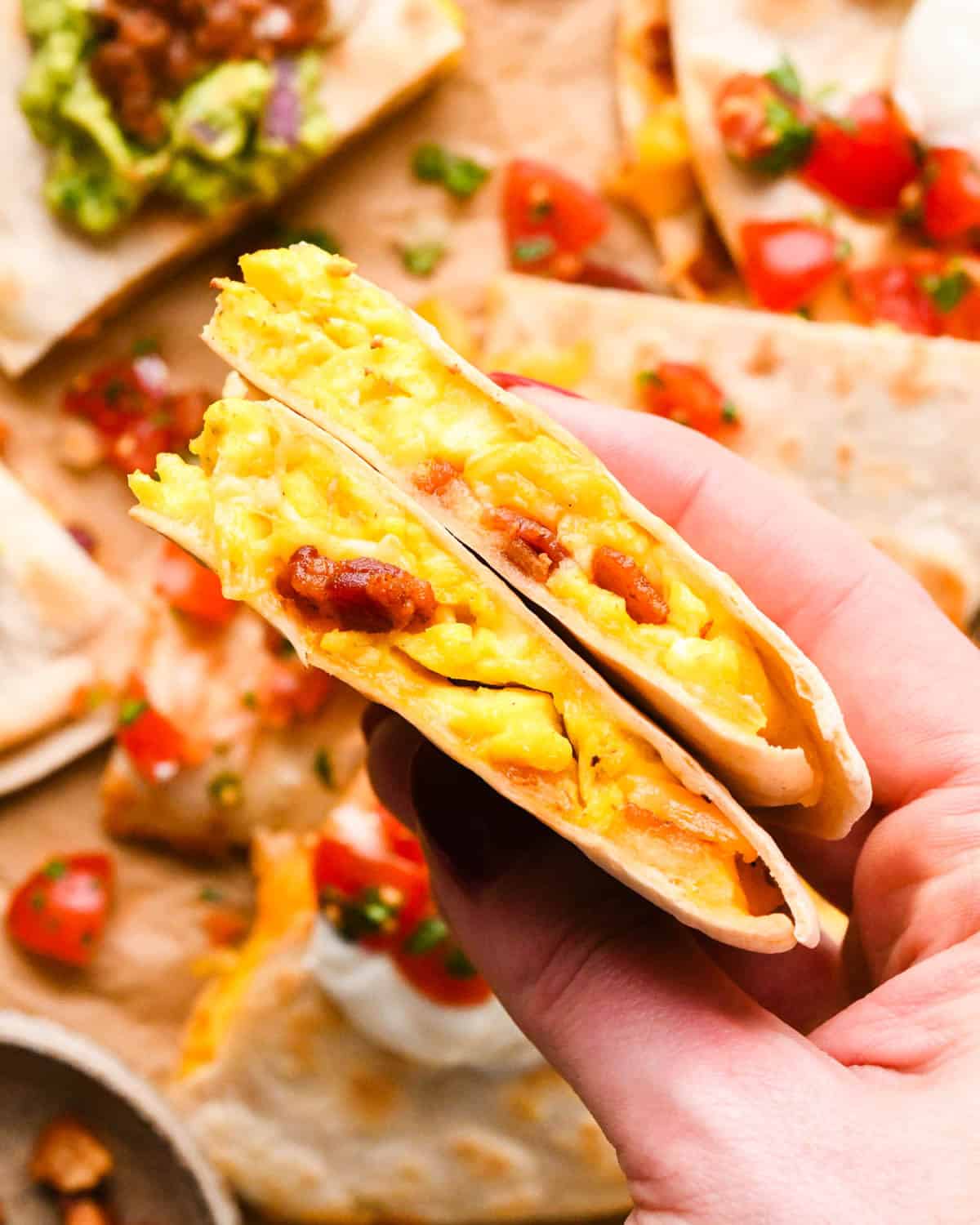 a hand holding two pieces of a breakfast quesadilla so the eggs, cheese and bacon inside are visible. It's hovering over more breakfast quesadillas topped with guacamole, sour cream and pico de gallo