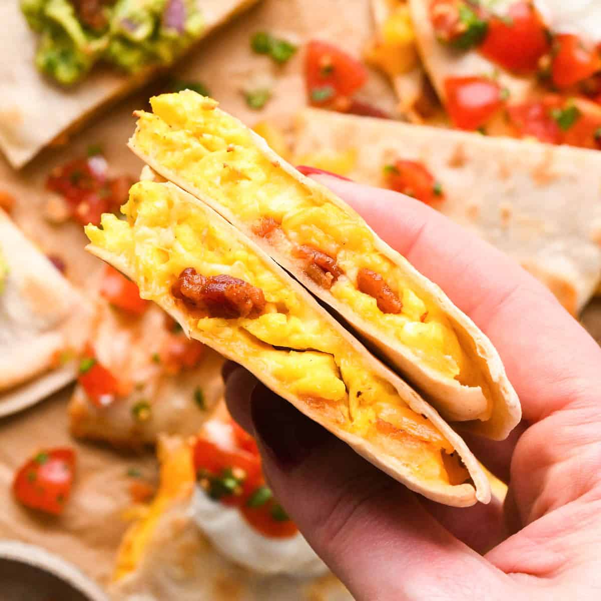 a hand holding two pieces of a breakfast quesadilla so the eggs, cheese and bacon inside are visible. It's hovering over more breakfast quesadillas topped with guacamole, sour cream and pico de gallo