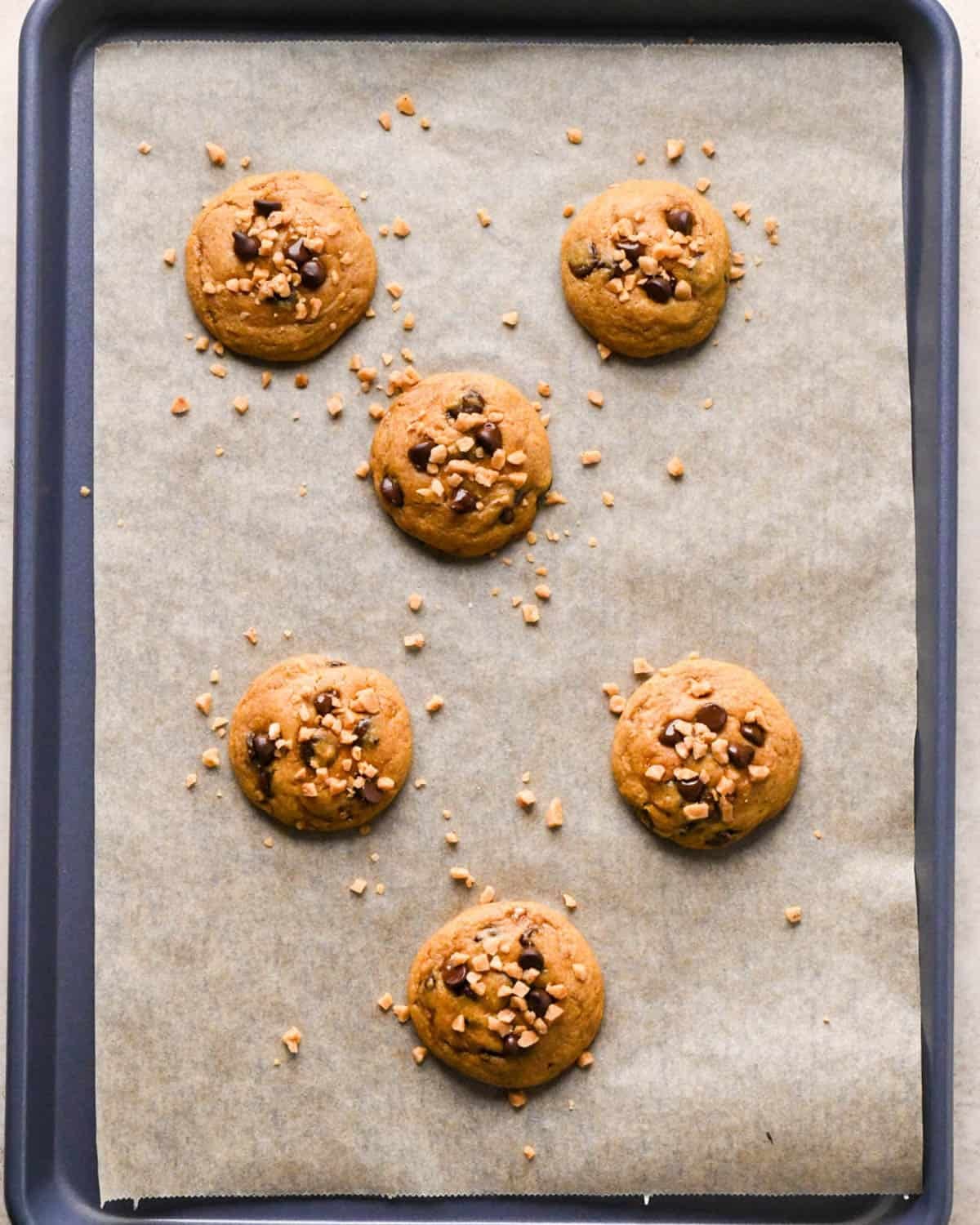 5 baked brown butter pumpkin cookies on a parchment lined baking sheet with extra chocolate chips and toffee bits added to the top