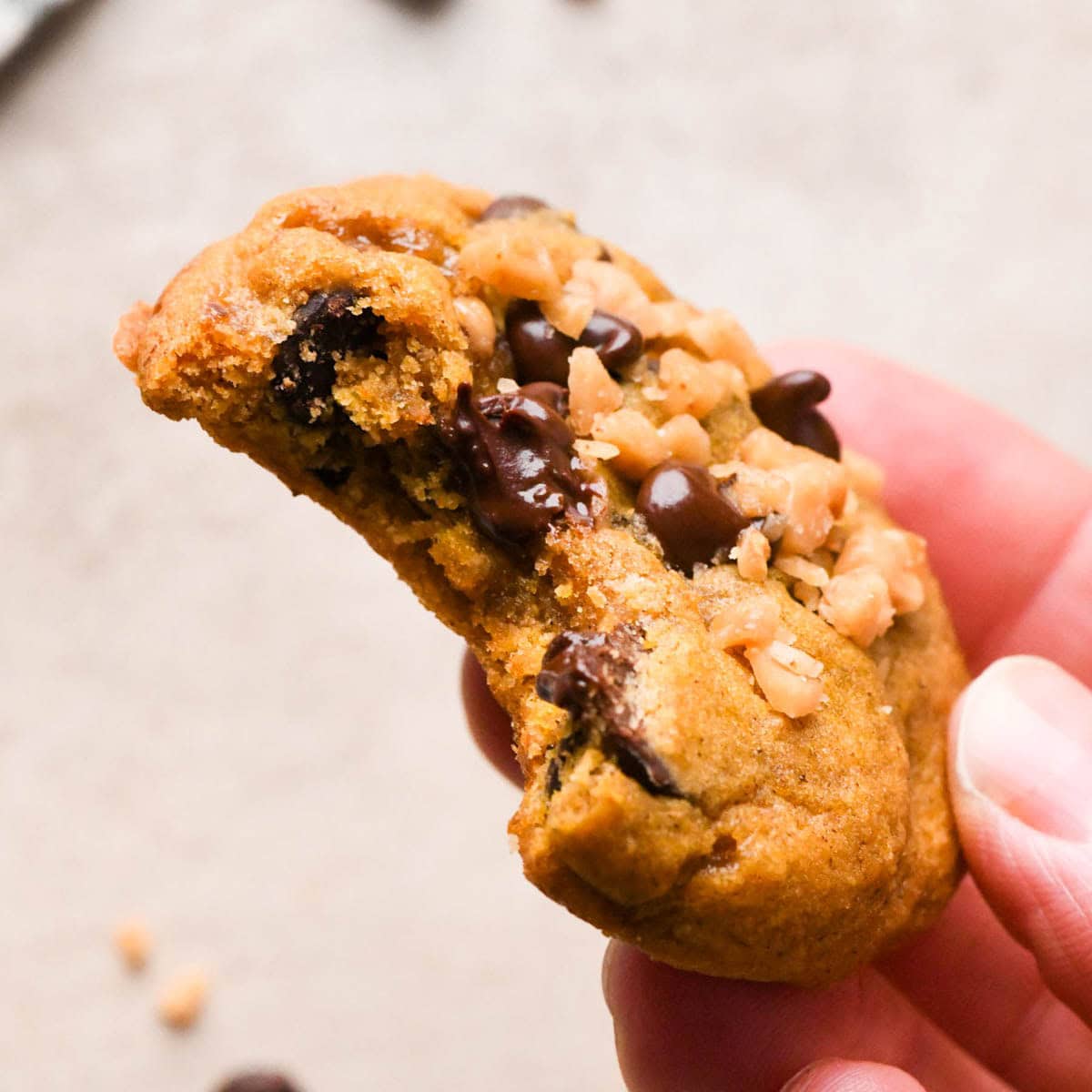 a hand holding a brown butter pumpkin cookie with a bite taken out of it, you can see melty chocolate chips and toffee bits on top