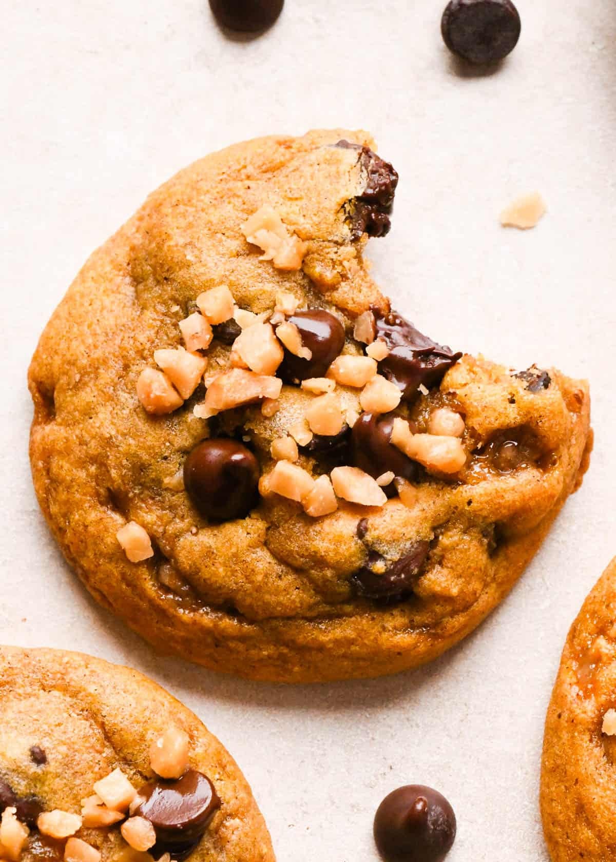 a brown butter pumpkin cookie with a bite taken out of it, with melty chocolate chips and toffee bits on top and two other cookies nearby