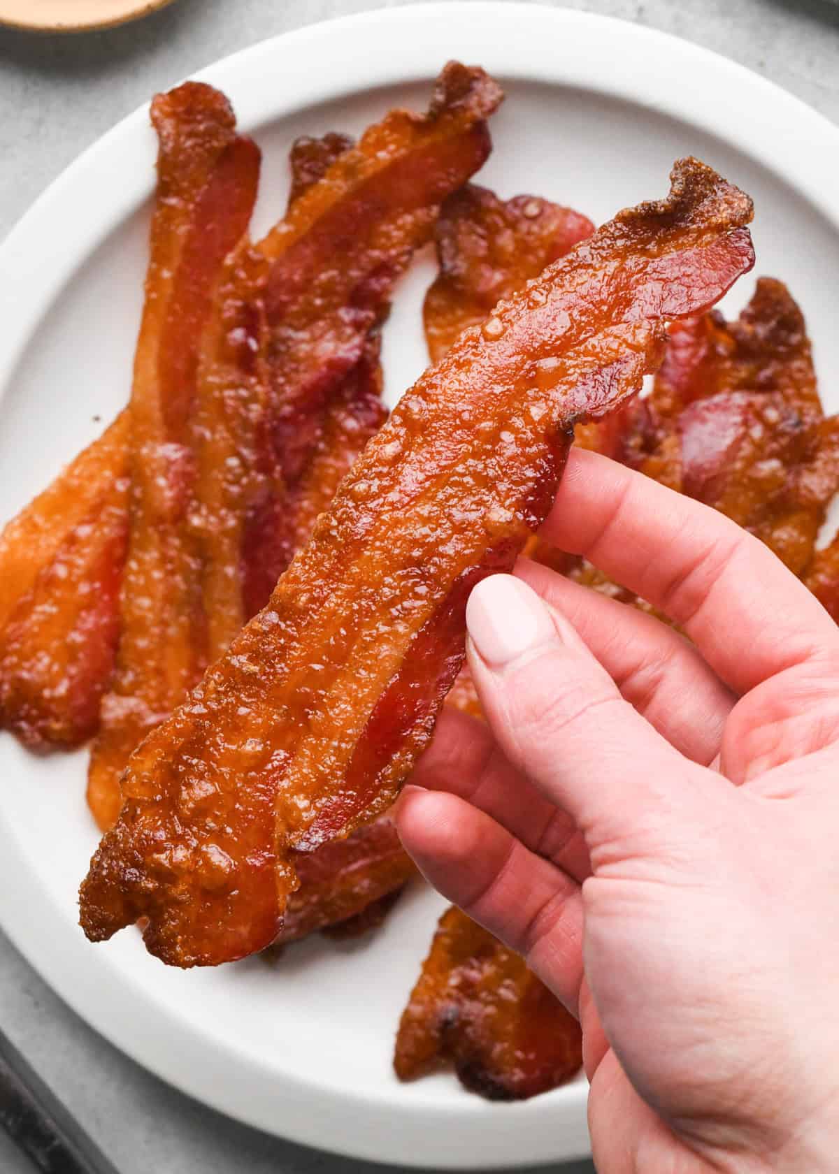 Overhead view of crispy brown sugar–glazed bacon strips arranged on a white plate on a light gray surface. A hand has picked up one of the bacon strips.