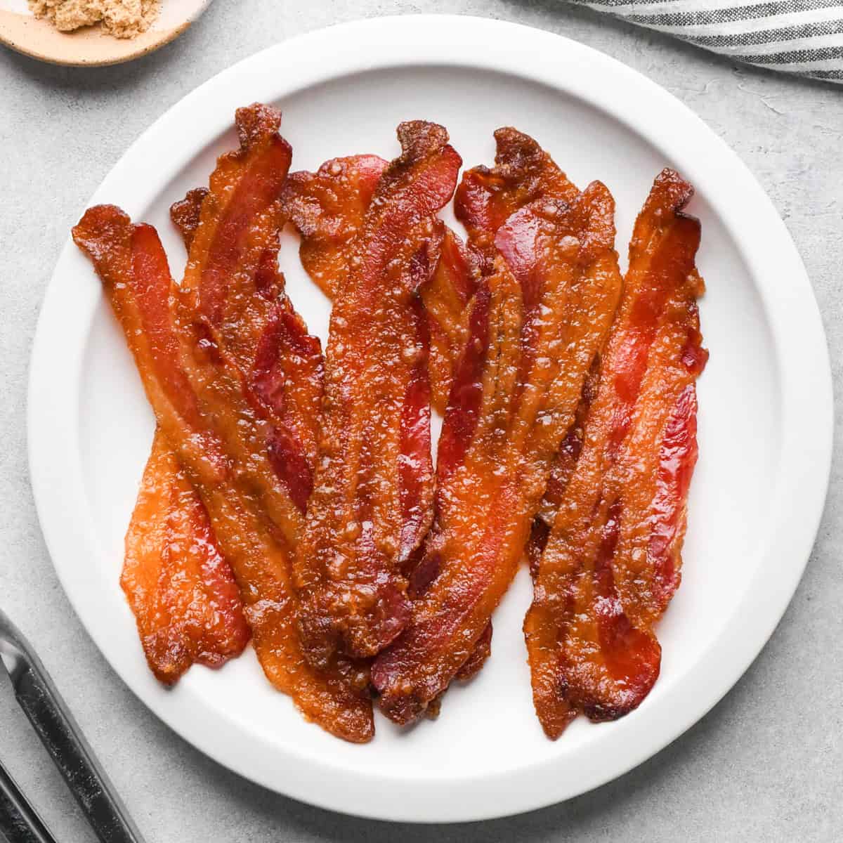 Overhead view of crispy brown sugar–glazed bacon strips arranged on a white plate on a light gray surface.