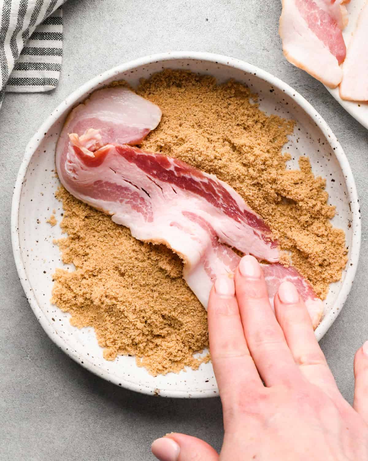 Making brown sugar bacon: overhead photo of a strip of bacon being coated in brown sugar on a white speckled plate.