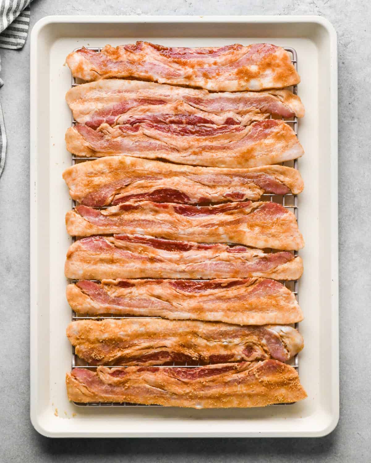 Making brown sugar bacon: overhead photo of 9 strips of brown sugar-coated bacon sitting on top of a cooling rack on a light-colored cooking sheet.