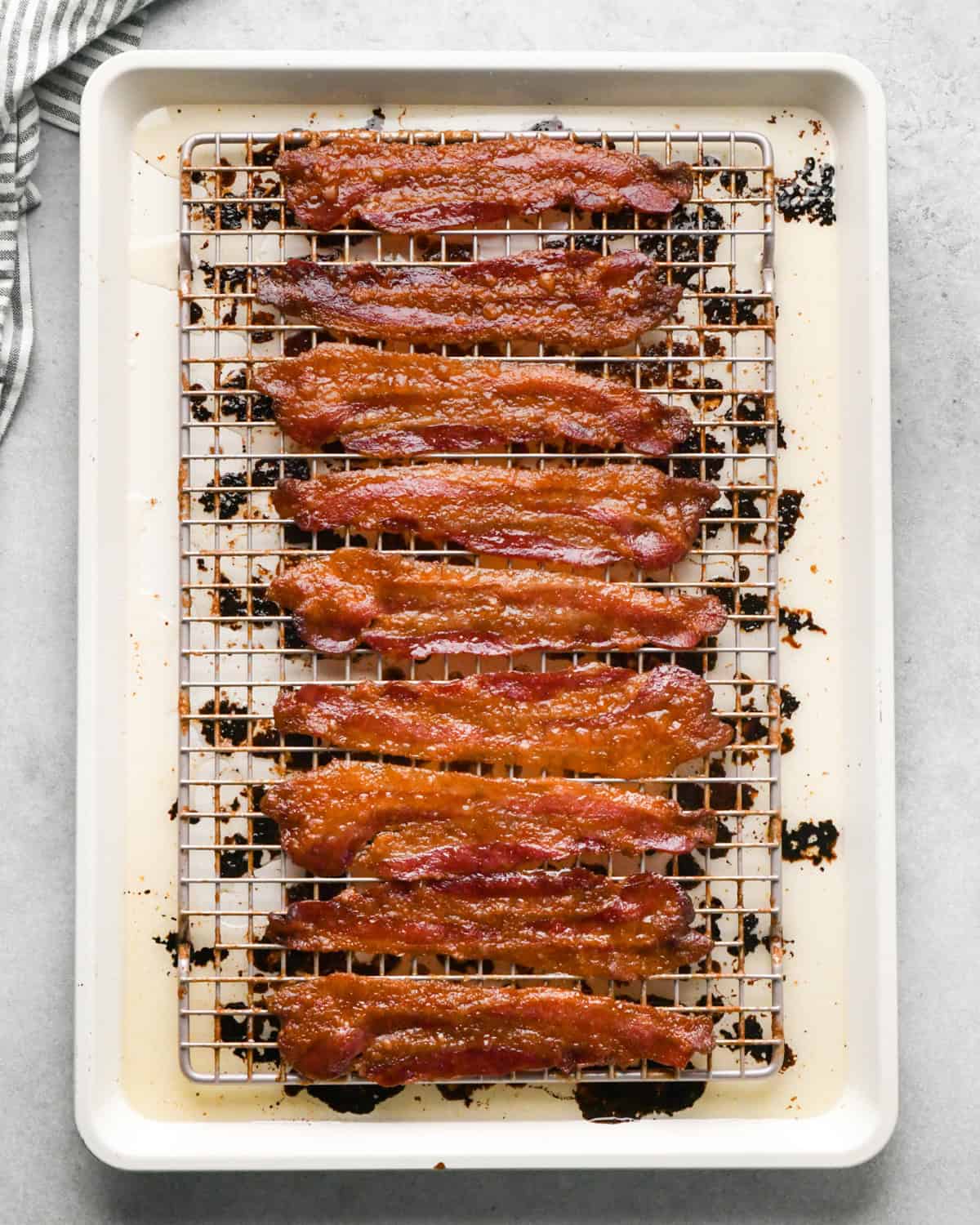 Making brown sugar bacon: overhead photo of 9 cooked strips of brown sugar-coated bacon sitting on top of a cooling rack on a light-colored cooking sheet.