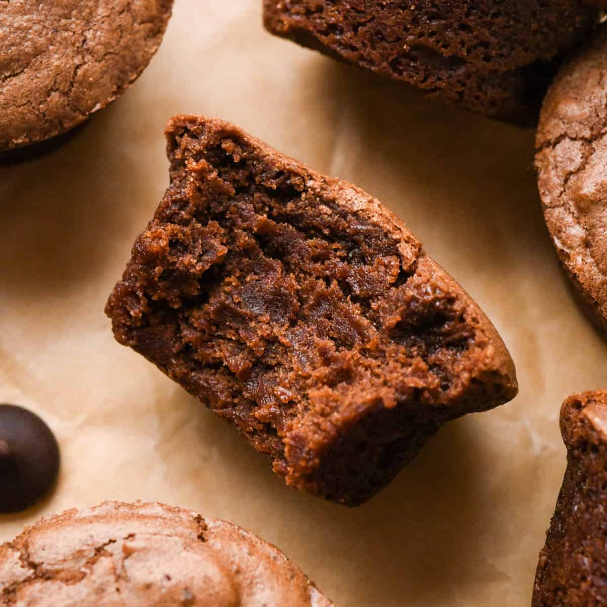 up close photo of a brownie bite with a bite taken out of it so the fudgy texture is showing, surrounded by other brownie bites and a chocolate chip all on brown parchment paper
