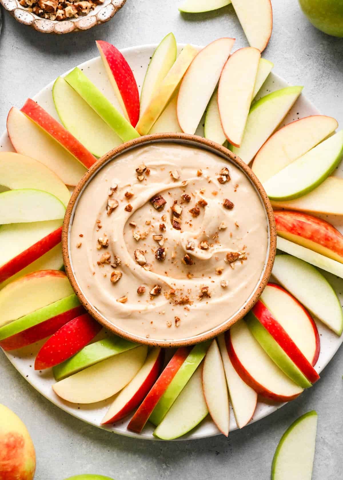 cream cheese apple dip in a pottery bowl with a brown rim topped with chopped pecans The bowl is on a plate with more red and green apple slices.