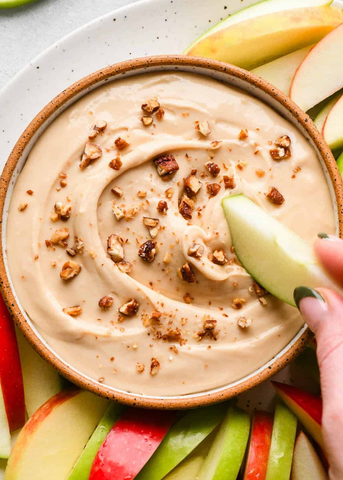 a green apple slice being dipped into a bowl of cream cheese apple dip surrounded by more red and green apple slices