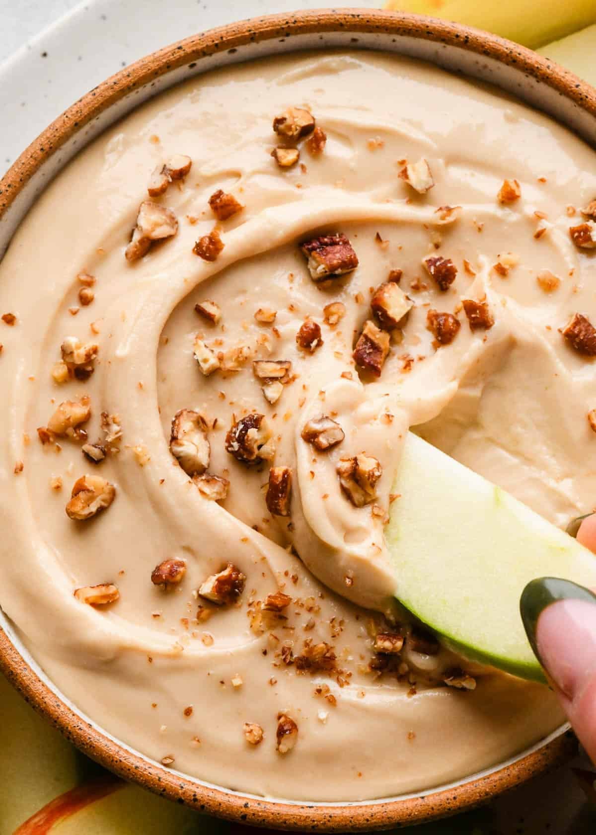 close up photo of a green apple slice being dipped into a bowl of cream cheese apple dip surrounded by more apples.
