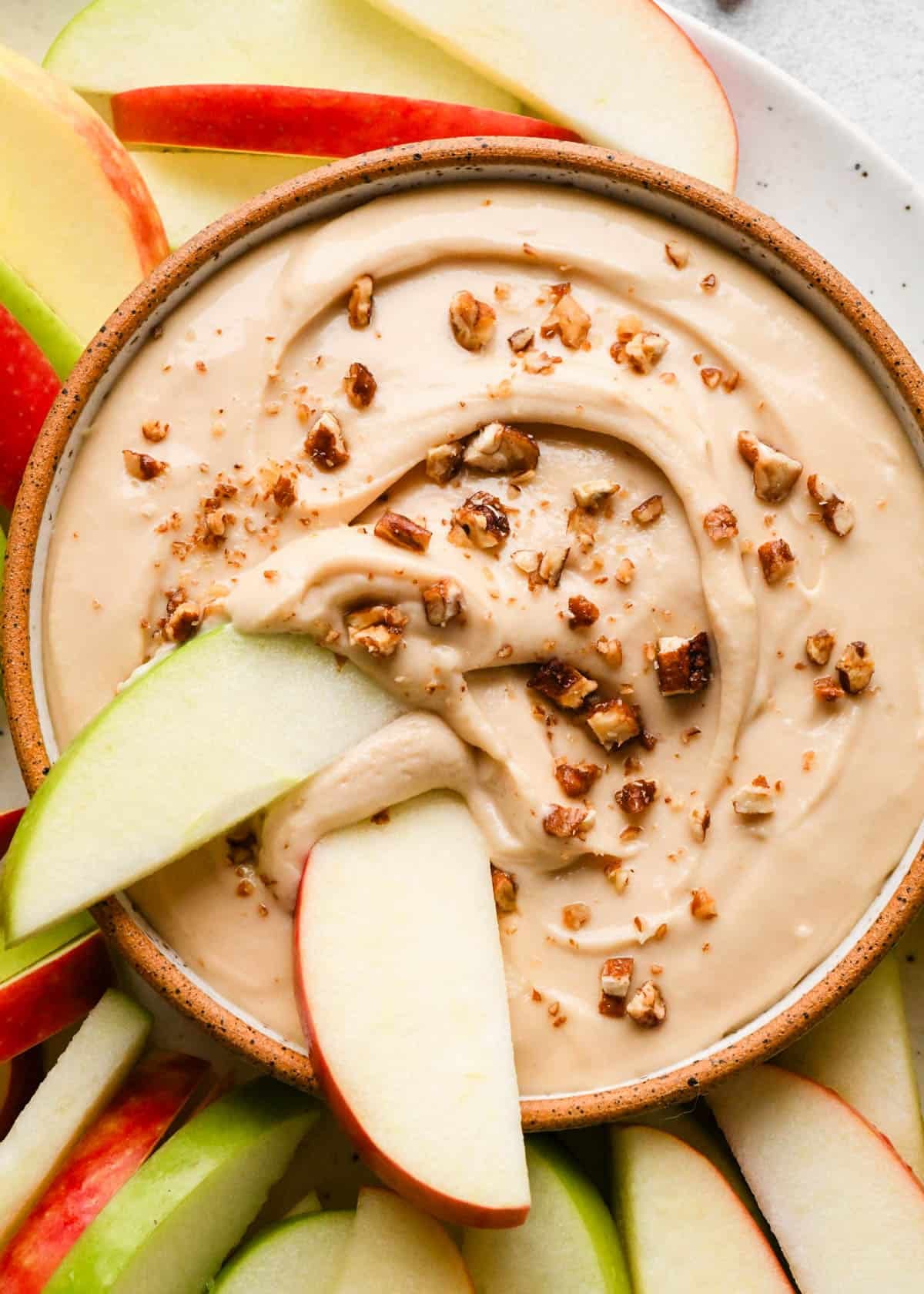 close up photo of cream cheese apple dip in a pottery bowl with a brown rim topped wit chopped pecans with two apple slices dipped in it, one red and one green. The bowl is on a plate with more red and green apple slices.